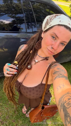 Woman with dreadlocks and tattoos wears a brown top, stands outdoors near a car.