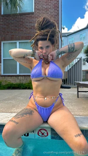 Woman with dreadlocks and tattoos in purple bikini sitting by pool, outdoor setting.
