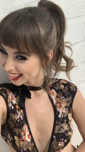 Woman with brown hair in ponytail wearing floral sheer top, posing indoors against white brick wall.