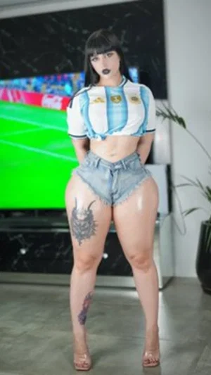 Woman in Argentina jersey and denim shorts, standing indoors with tattoos on legs.