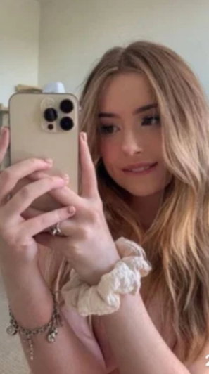Woman with long blonde hair taking mirror selfie, wearing light pink top, indoors.