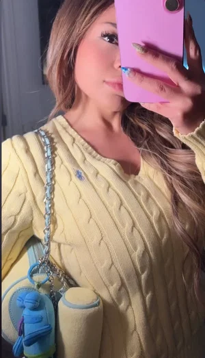 Woman with long hair in yellow sweater holding pink phone, wearing blue purse, indoor setting.