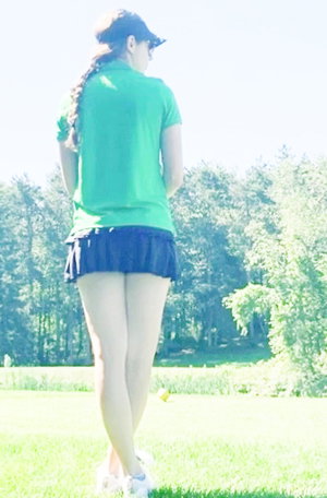 Woman in green shirt and blue skirt standing outdoors on grass, back view.