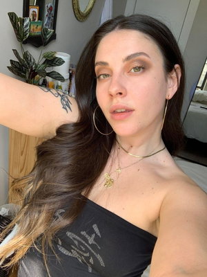 Woman with long dark hair in black strapless top, taking selfie in bedroom.