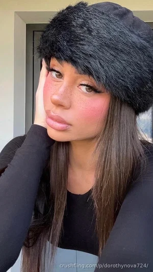 Woman with long brown hair wearing black fur hat and black top, posing indoors.