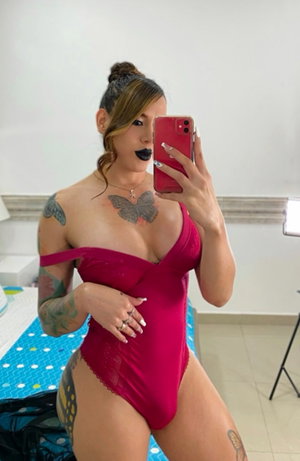 Tattooed woman in red lace bodysuit, bedroom selfie.
