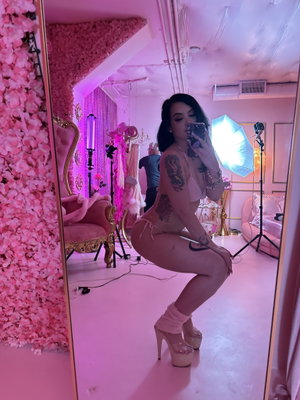 Tattooed woman in pink lingerie posing for mirror selfie in studio.