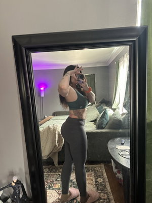 Woman in sports bra and leggings taking mirror selfie in bedroom.