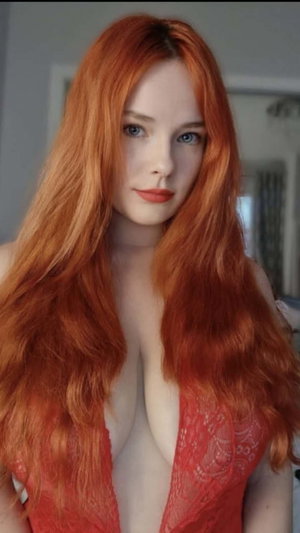 Woman with long red hair in red lace top, indoors, facing camera.