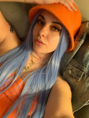 Woman with blue hair and orange hat, wearing orange top, lying on couch with denim jacket.