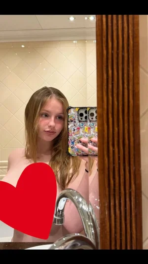 Woman with wet hair taking mirror selfie in bathroom, topless with red heart overlay.