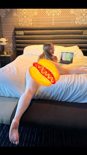 Woman with blonde hair sits on bed with laptop, wearing inflatable hot dog costume.