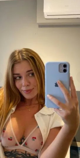 Woman with blonde hair in a floral bra and open white shirt, taking mirror selfie indoors.