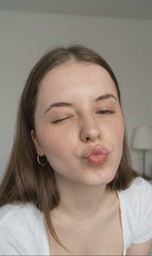 Woman with brown hair in white top winking and puckering lips indoors.
