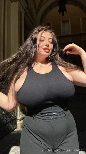 Woman with long dark hair in black tank top and pants, posing outdoors in a sunlit alley.