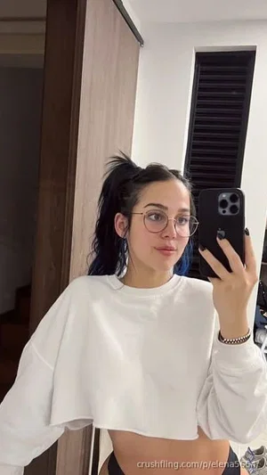 Woman with blue-tipped hair and glasses wears white crop top, holding phone for mirror selfie in