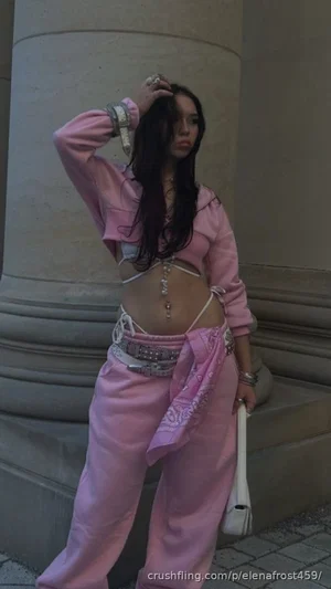 Woman in pink outfit with crop top and pants, standing near stone column outdoors.