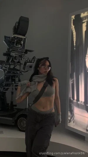 Woman in gray crop top and cargo pants, wearing glasses, standing near camera equipment indoors.