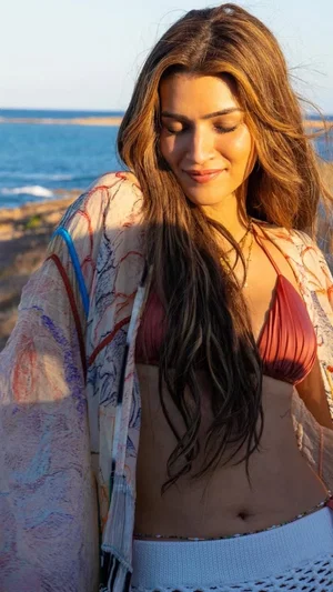 Woman with long brown hair in red bikini top and white skirt, standing by ocean.
