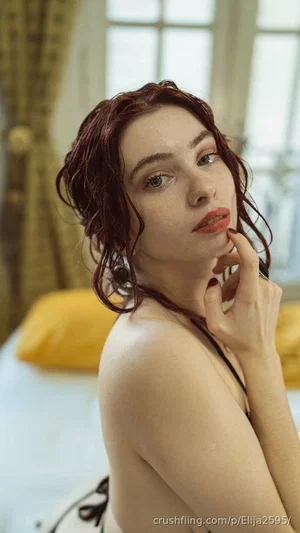 Woman with wet red hair in a bedroom, wearing a black strap top, touching her face.