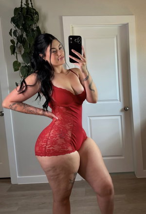 Woman in red lace bodysuit posing in indoor setting with tattoos visible.