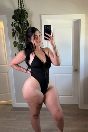 Woman in black bodysuit poses for mirror selfie in indoor setting with tattoos visible.