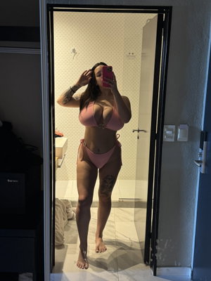 Woman in pink bikini taking mirror selfie in tiled bathroom.