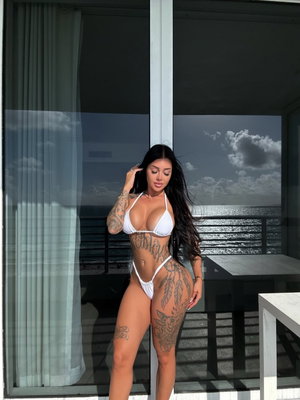 Woman in white bikini with tattoos poses on balcony overlooking ocean.