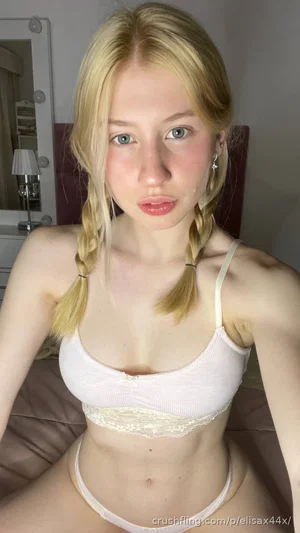 Blonde woman in white lingerie, sitting on bed, mirror selfie, indoor setting.