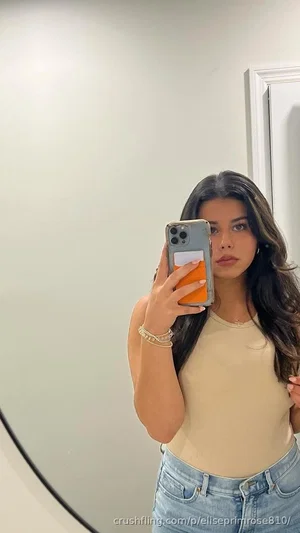 Woman with long dark hair in beige top and jeans taking mirror selfie indoors.