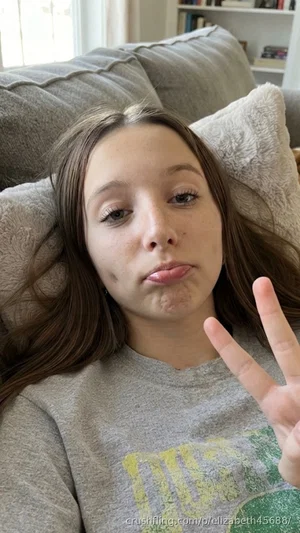 Woman with long brown hair in gray sweatshirt lying on couch, making peace sign.
