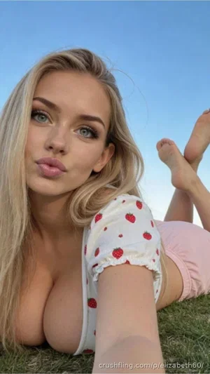 Blonde woman in strawberry-patterned top and pink shorts lying on grass, making a playful face.