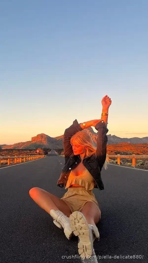 Woman with blonde hair sits on road, wearing crop top, skirt, jacket, and boots, stretching arms,