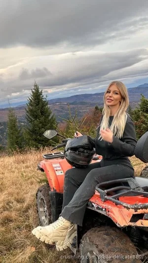 Woman with blonde hair in black outfit sitting on orange ATV in grassy outdoor setting with