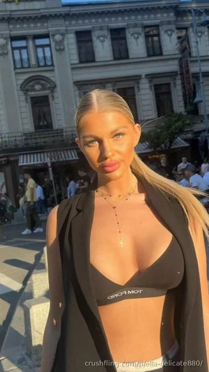 Woman with blonde hair in black bra and blazer, standing outdoors in city setting.
