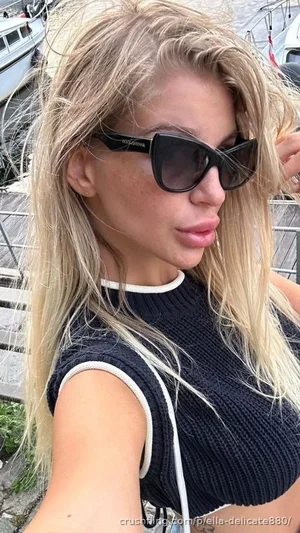 Blonde woman in black crop top and sunglasses, outdoors near boats.
