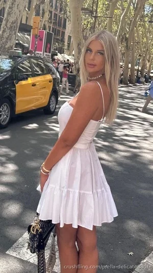 Woman in white dress posing on city street with trees and cars.