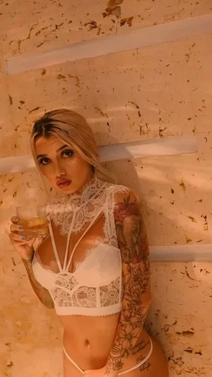 Woman with blonde hair and tattoos wears white lace lingerie, holding a drink against a textured