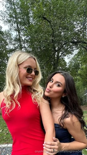 Two women in dresses posing outdoors, one with blonde hair, one with dark hair, smiling and playful.