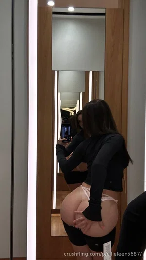 Woman in black crop top and underwear posing in mirror selfie, showing backside.