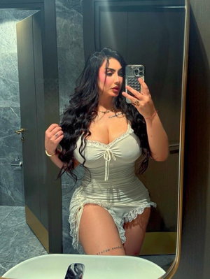 Woman with long dark hair in light-colored lace lingerie taking mirror selfie in bathroom.