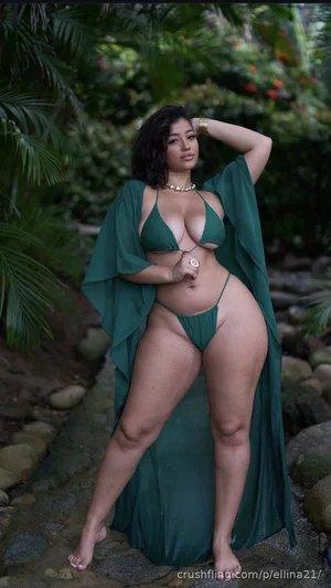 Woman in green bikini and sheer robe posing outdoors near rocks and plants.