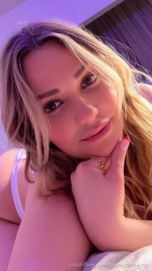 Woman with blonde hair in white top posing indoors, hand on chin.