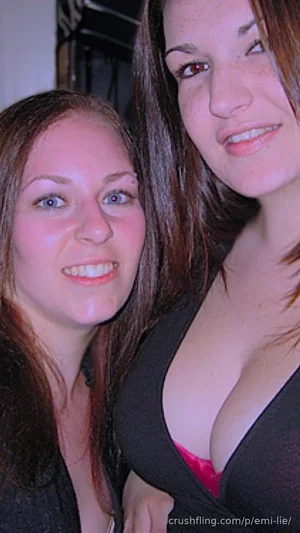 Two women with long brown hair, one in a black top with pink lining, posing closely indoors.