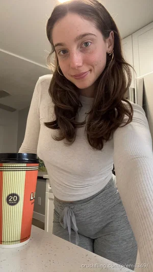 Woman with long brown hair in a white long-sleeve top and gray sweatpants, standing in a kitchen.