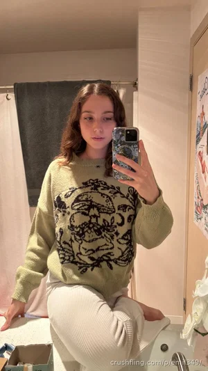 Woman in olive sweater with mushroom design, white pants, sitting on bathroom counter, mirror
