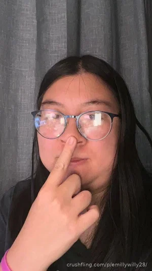 Woman with long dark hair and glasses, wearing black top, touching nose, gray curtain background.