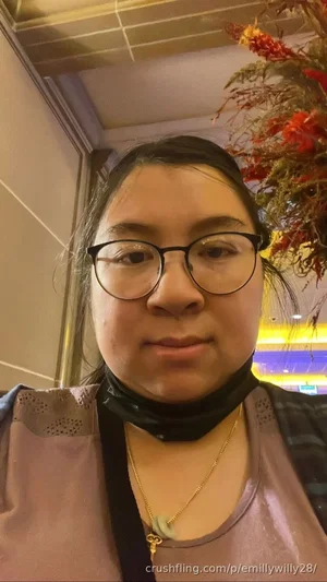 Woman with glasses, dark hair, wearing a purple top and gold necklace, indoors with floral decor.