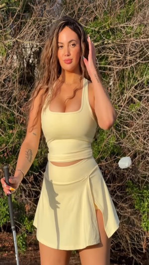 Curvy woman in yellow tank top and skirt holds golf club outdoors.