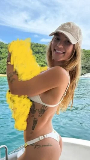 Woman in white bikini and cap on boat, holding yellow object, tattoos visible, smiling.